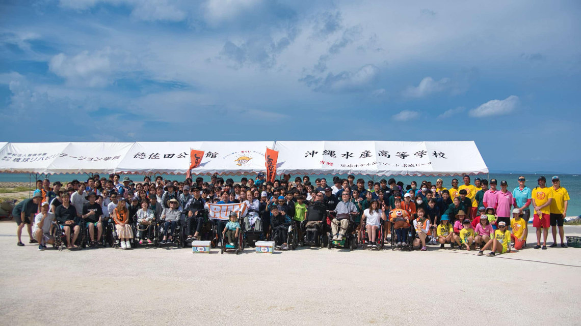Large group of people, including wheelchair users, pose together under a tent at Yuimaru Beach Festival by the ocean.