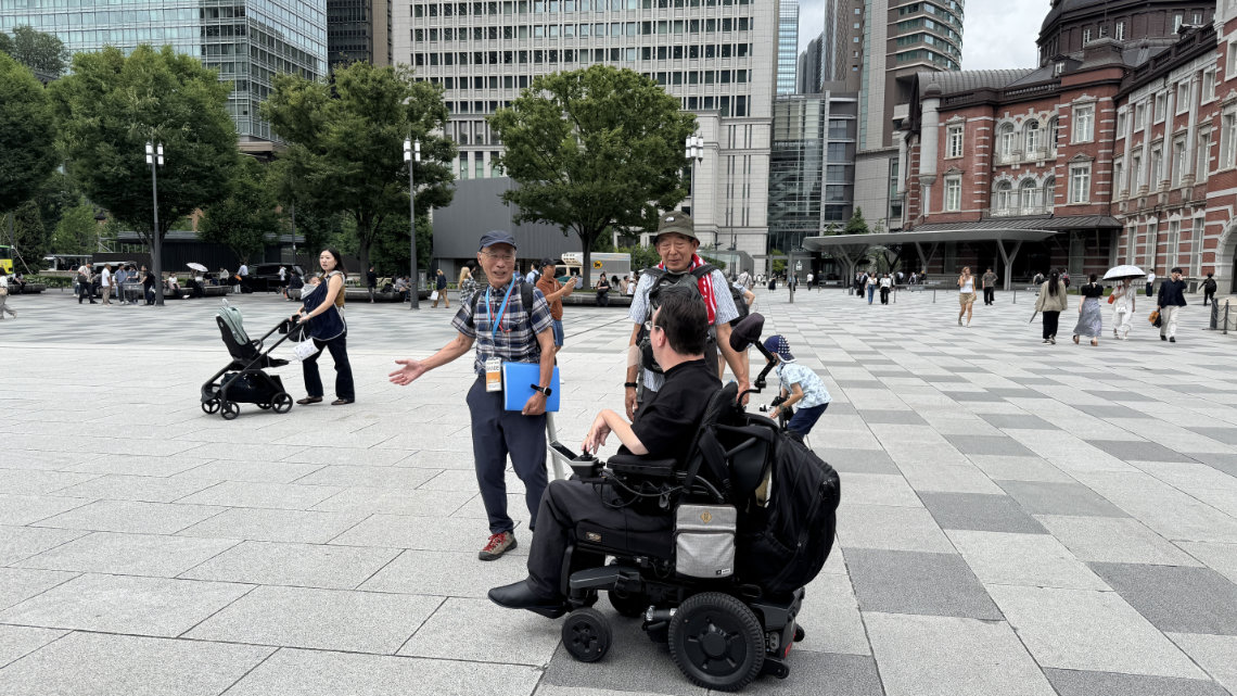A man in a wheelchair is guided by volunteer tour guides near Tokyo Station, with people and city buildings in the background.