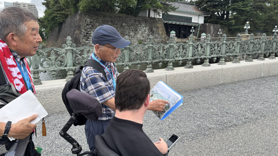 Volunteer guide shows a map to a wheelchair user near the Imperial Palace bridge and stone moat wall.