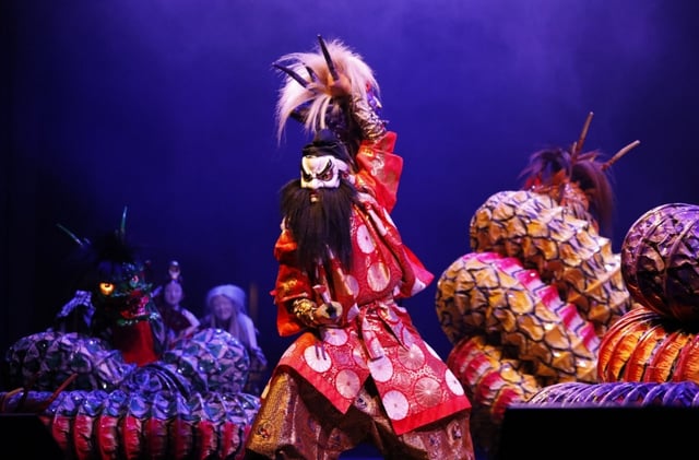 A performer in a vibrant red costume and mask raises a sword during the traditional Japanese dance-drama Iwami Kagura, with dragon figures in the background on stage.