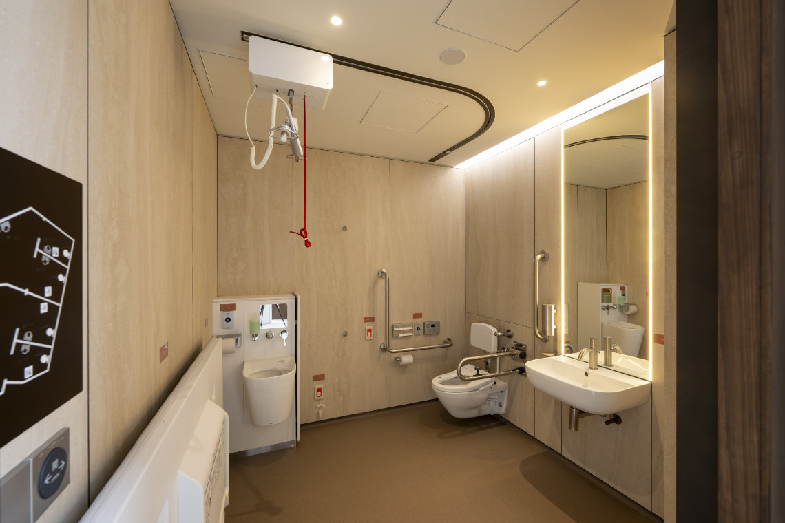 Accessible restroom with support rails, ceiling lift, baby changing unit, and modern sink under warm lighting.
