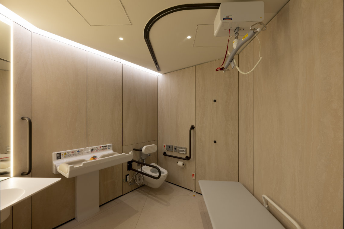 Spacious accessible restroom with a ceiling lift, wall-mounted toilet, baby changing table, and padded bench.