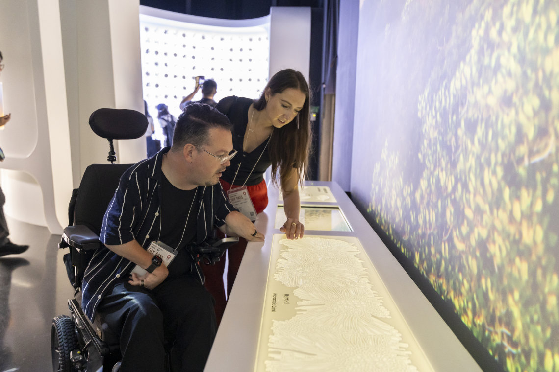 A man in a wheelchair and a woman examine a tactile display inside the Saudi Arabia Pavilion at Expo 2025.