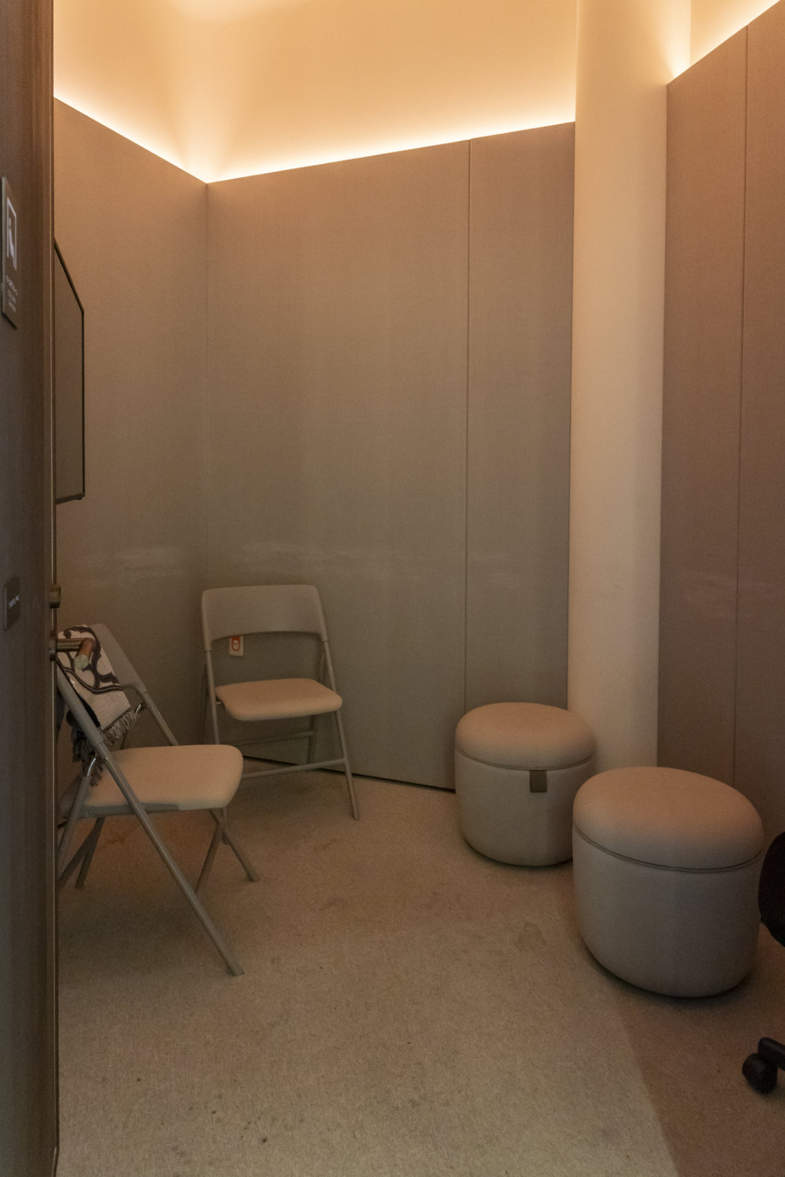 Quiet room with soft lighting, two folding chairs, and two round cushioned stools against beige walls.