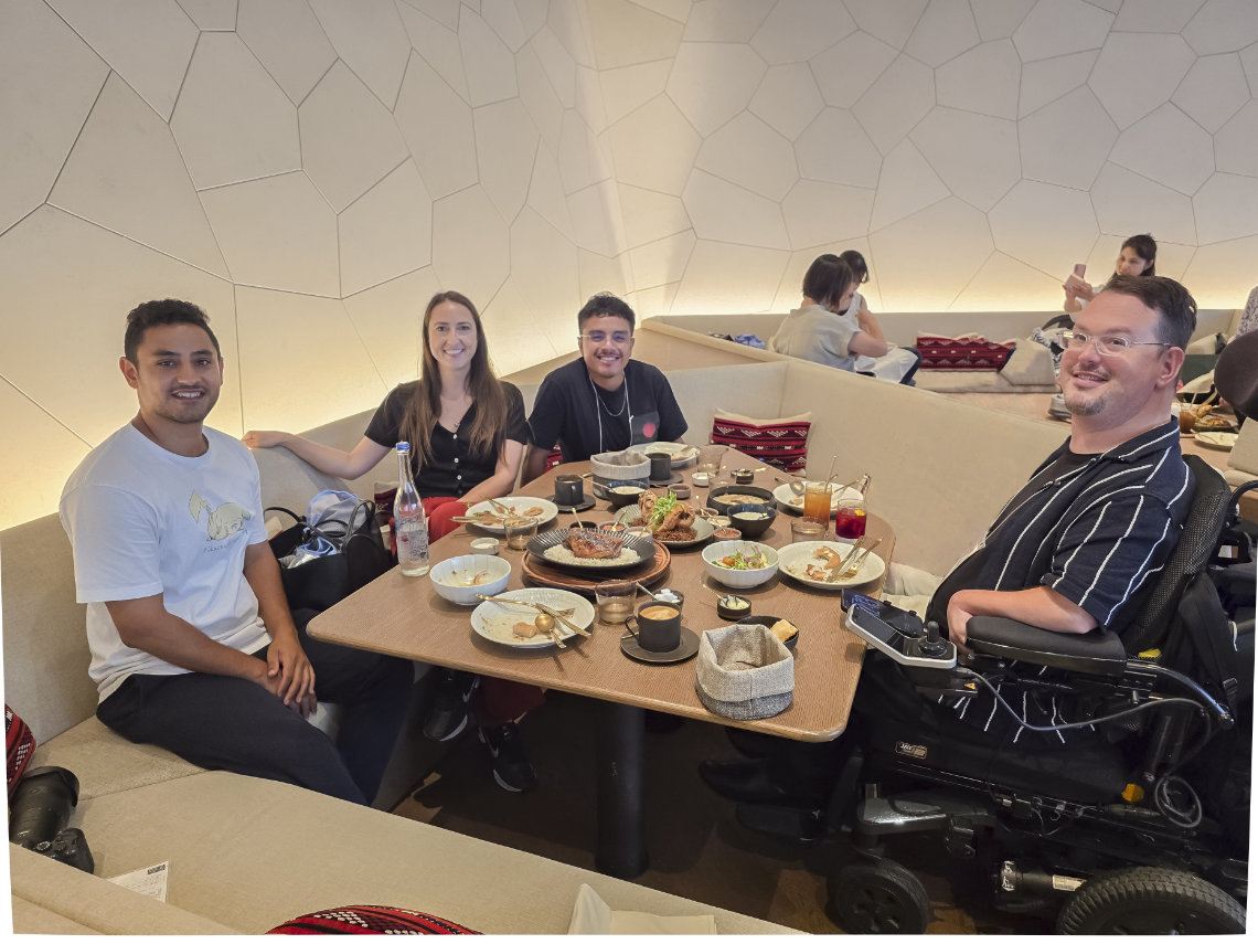 Four friends sit around a restaurant table enjoying a meal, smiling amid plates of food and drinks.