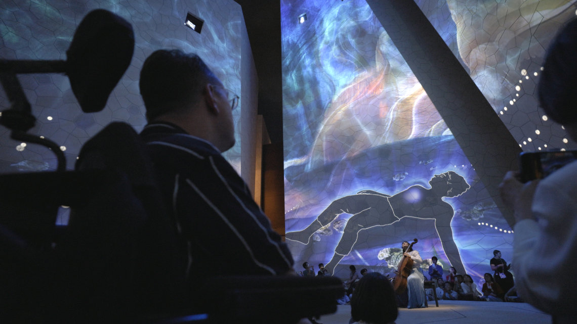 Audience watches a live cello performance beneath vivid, immersive projections on tall geometric walls.