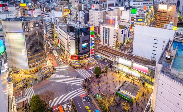 A vibrant aerial view of the famous Shibuya Crossing in Tokyo, Japan, taken during the evening. Crowds of people are seen crossing the wide intersection from all directions beneath the glow of colorful billboards and neon signs. Surrounding the crossing are tall buildings, shops, and restaurants, while a train passes on elevated tracks nearby. The energy of the city is captured with bustling streets, bright lights, and a lively urban atmosphere.