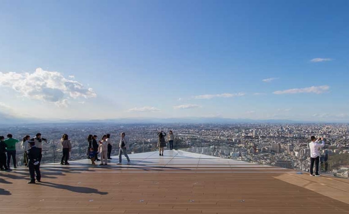 Visitors stand on the open-air observation deck of Shibuya Sky in Tokyo, Japan, enjoying panoramic views of the city under a clear blue sky. The wooden deck and glass barriers create a modern, spacious atmosphere, while people take photos and admire the skyline stretching out to the horizon.