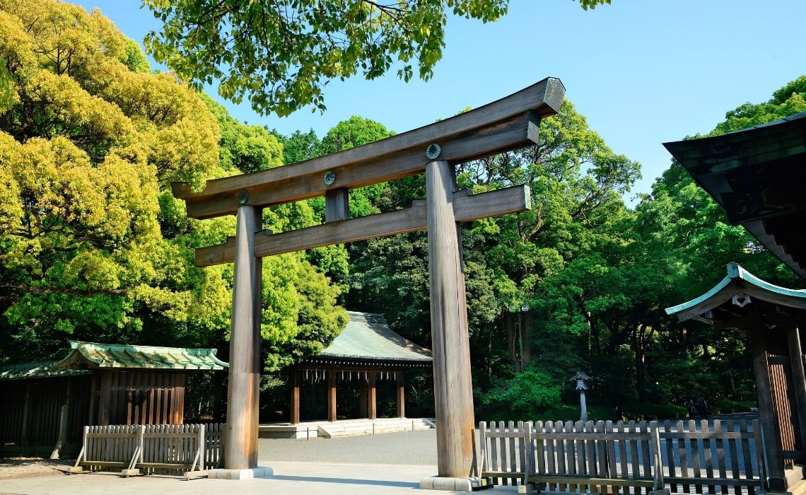 A large traditional wooden torii gate stands at the entrance of Meiji Shrine in Tokyo, Japan, surrounded by lush green trees and peaceful natural scenery. The sunlight filters through the leaves, illuminating the pathway leading toward the shrine buildings in the background.