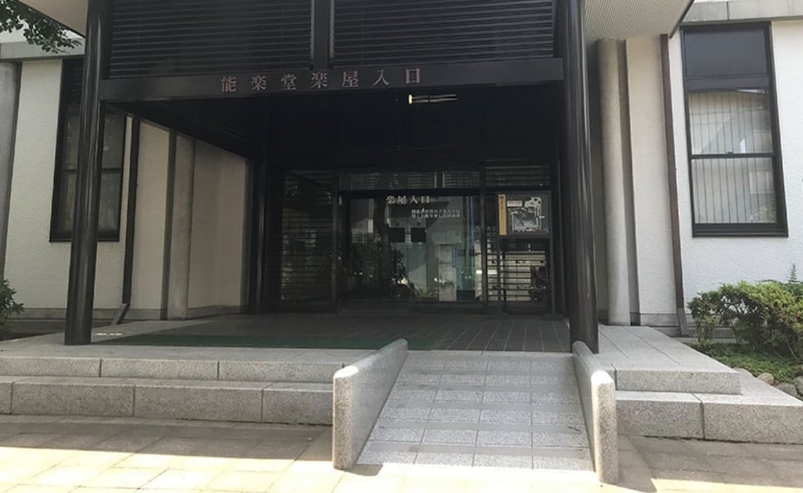 An accessible entrance to a building in Japan, featuring a smooth concrete ramp with raised edges leading up to a set of glass doors. The entrance is shaded by a black metal awning supported by columns, with signage in Japanese above the doorway and small bushes lining the sides.
