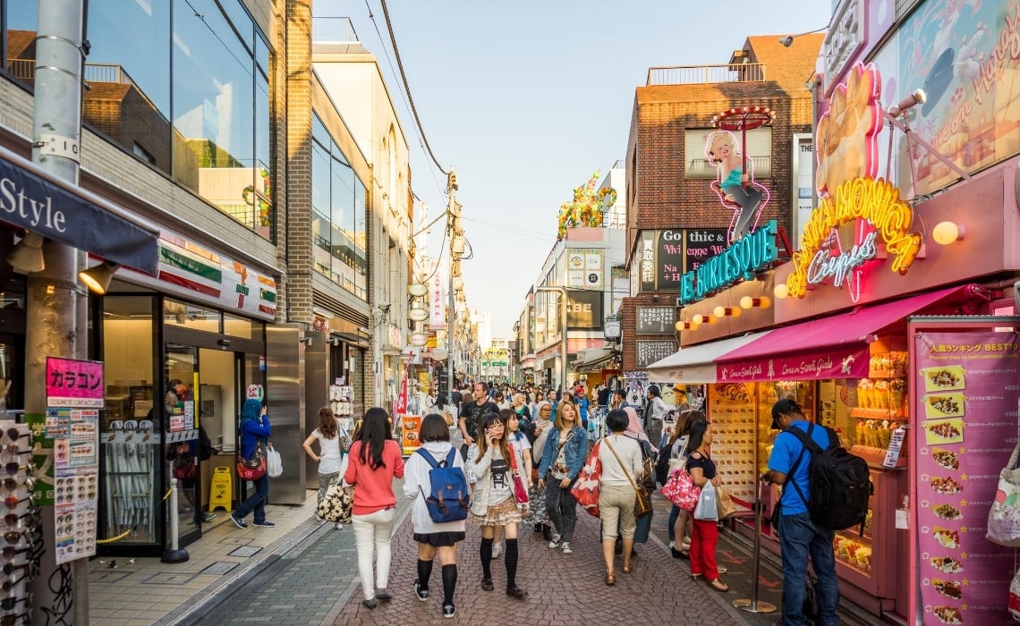 A lively pedestrian street in Harajuku, Tokyo, filled with people shopping and strolling among colorful storefronts and signs. On the right, a bright pink crepe shop with neon signage adds to the playful atmosphere, while other shops line the narrow street under a clear afternoon sky. The scene captures the youthful and trendy energy of Harajuku’s Takeshita Street.