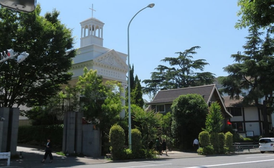 A white church with a tall bell tower topped by a cross stands among lush green trees on a sunny day in Tokyo. The building features classical architectural details, while nearby smaller structures and people walking along the street add to the calm, suburban atmosphere.
