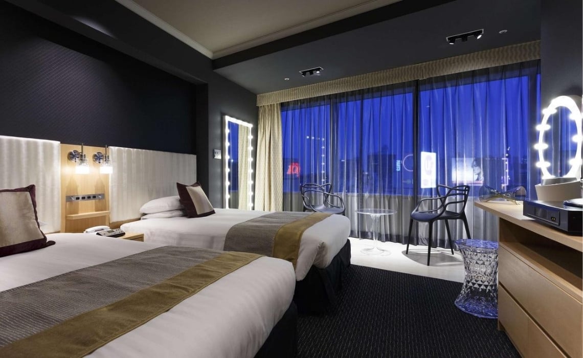 A modern twin hotel room featuring two neatly made beds with gold and gray runners, elegant lighting, and a large window with sheer curtains offering a view of Tokyo’s illuminated skyline at night. The room includes a vanity mirror surrounded by lights, contemporary chairs, and a small glass table, creating a stylish and comfortable atmosphere.