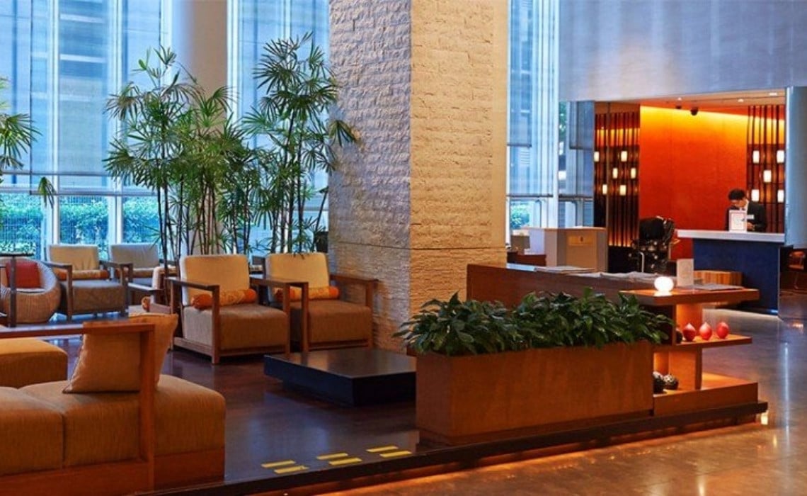 A modern hotel lobby featuring warm lighting, wooden furniture with beige and orange upholstery, and large indoor plants adding a natural touch. The space includes comfortable seating areas, a stone pillar at the center, and a reception desk in the background where a staff member assists a guest. Tall windows let in daylight, creating an inviting and elegant atmosphere.