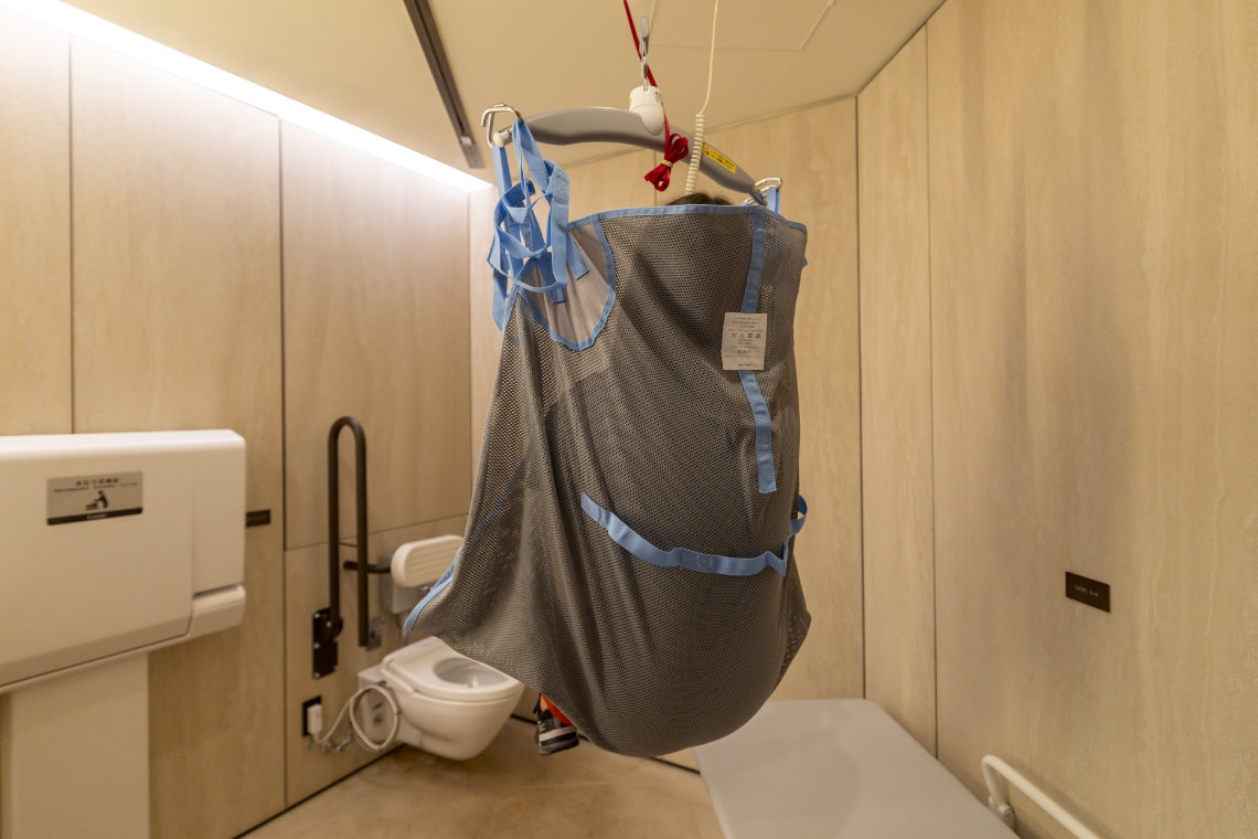Ceiling lift with a gray and blue sling suspended above an accessible restroom with beige walls.