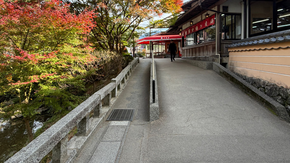 Gentle uphill slope by shops and autumn trees near Dazaifu Tenmangu, with a handrail and accessible path beside a stream.