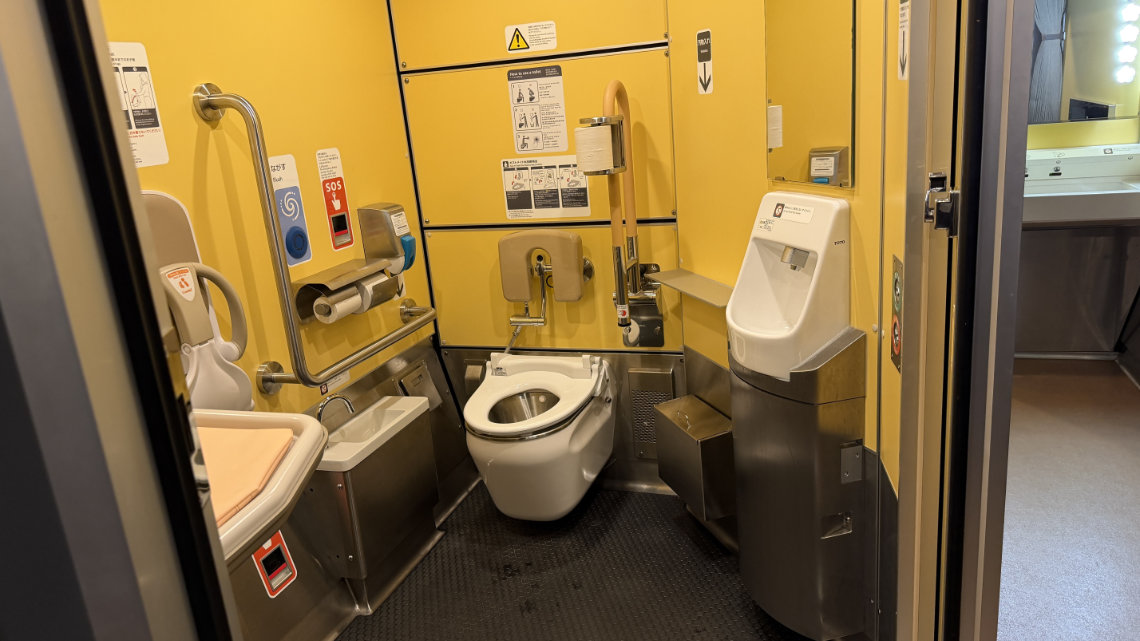A spacious, yellow-walled accessible train restroom equipped with a toilet with grab bars, a urinal, a lower sink, and a wall-mounted baby seat.