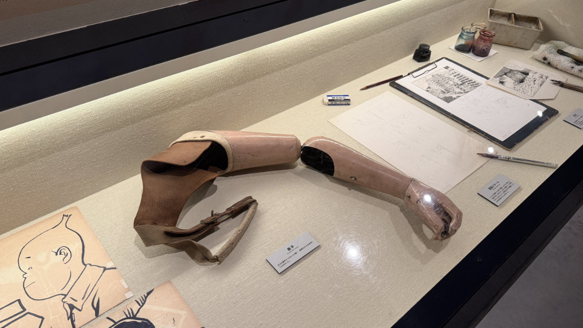 A museum glass case displaying a vintage prosthetic arm alongside manga sketches, ink bottles, and drawing pens arranged on a light fabric surface.