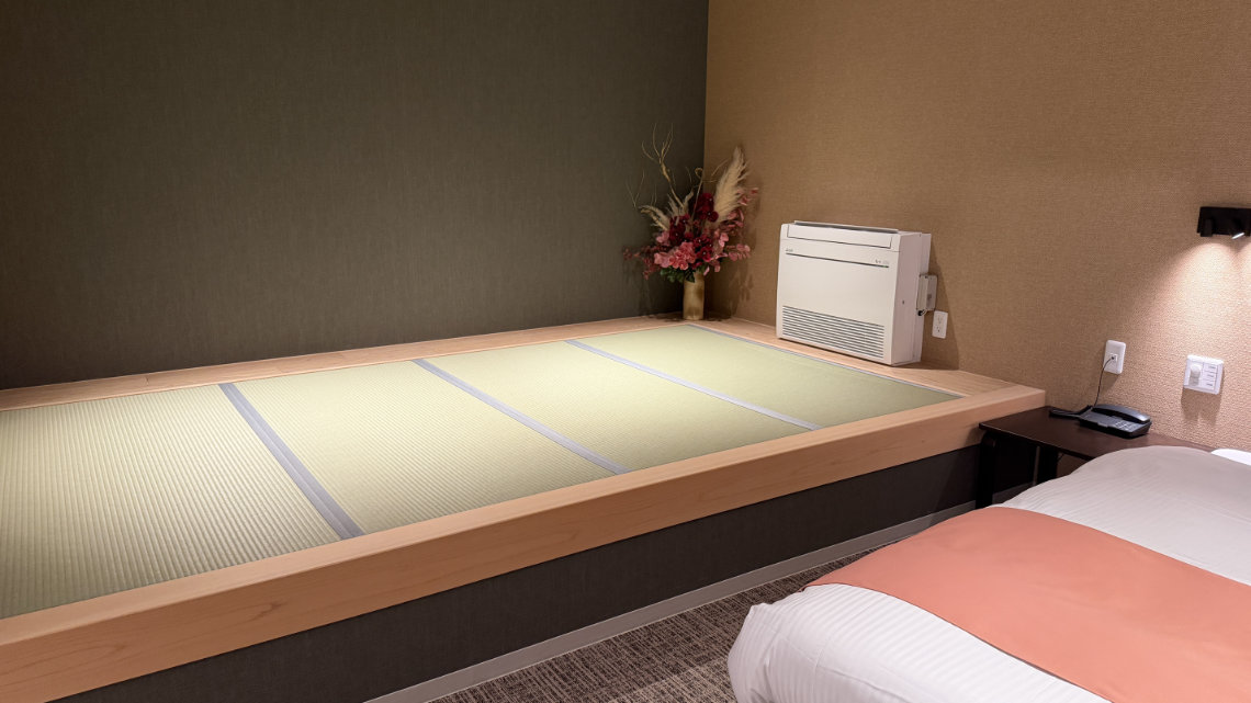 A hotel room featuring a twin bed next to a raised wooden platform containing tatami mats. A white heater and a flower arrangement sit on the platform against the wall.