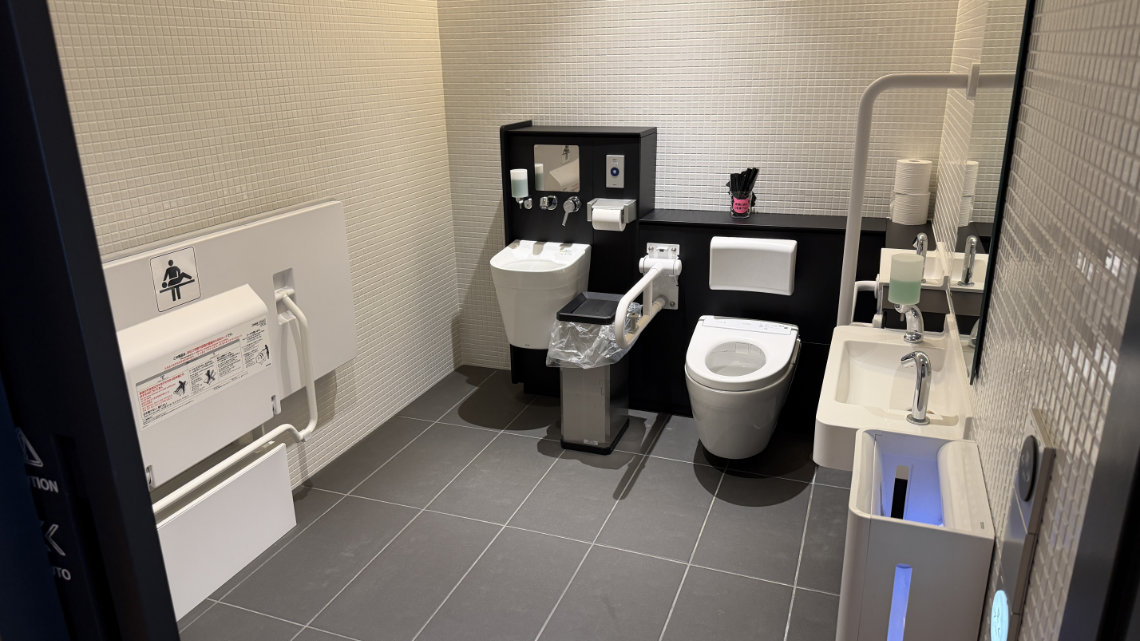A fully equipped accessible restroom with white tiled walls, featuring a toilet with grab bars, a washbasin, and a large fold-down changing bed on the left wall.