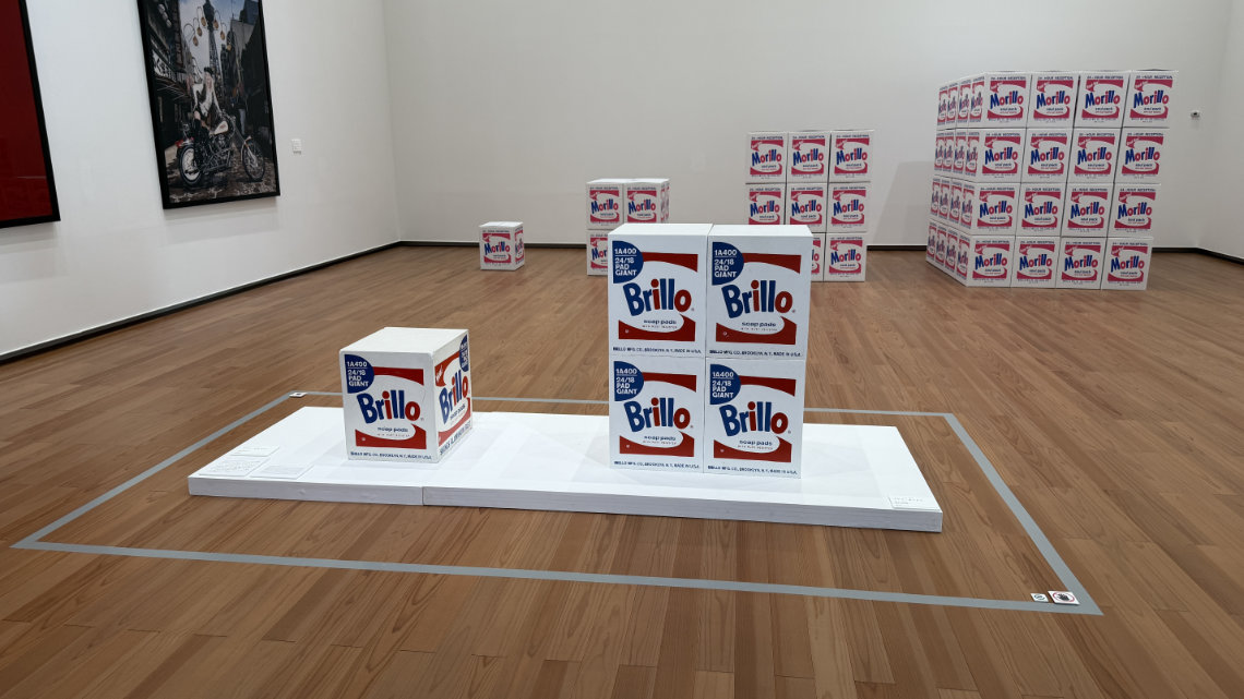 A museum gallery with wood floors displaying stacks of white "Brillo Soap Pads" boxes on a low platform, with other box art installations in the background.