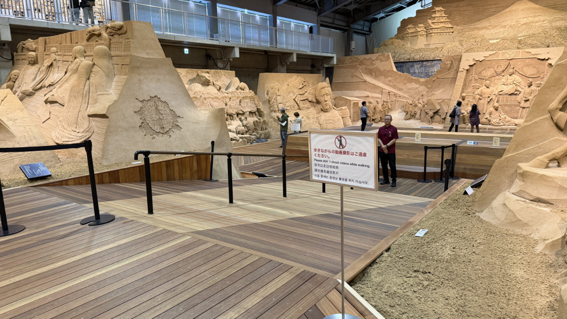 Large elaborate sand sculptures viewed from a wooden ramp, with a sign in the foreground warning against shooting video while walking.