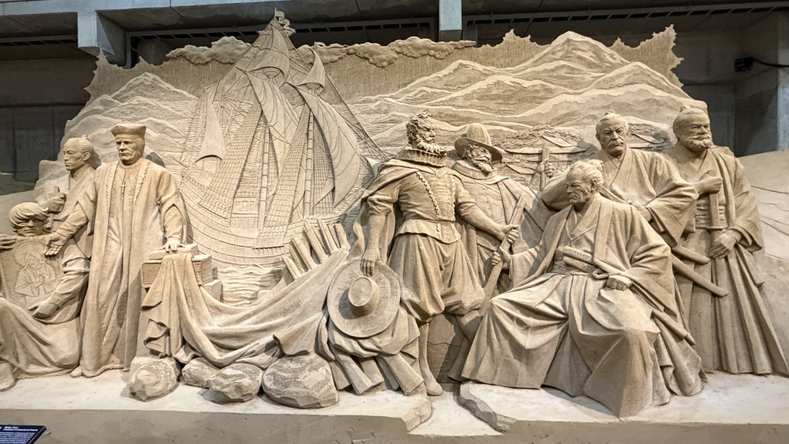 A detailed sand sculpture relief depicting historical figures, including samurai and European missionaries, alongside a sailing ship.
