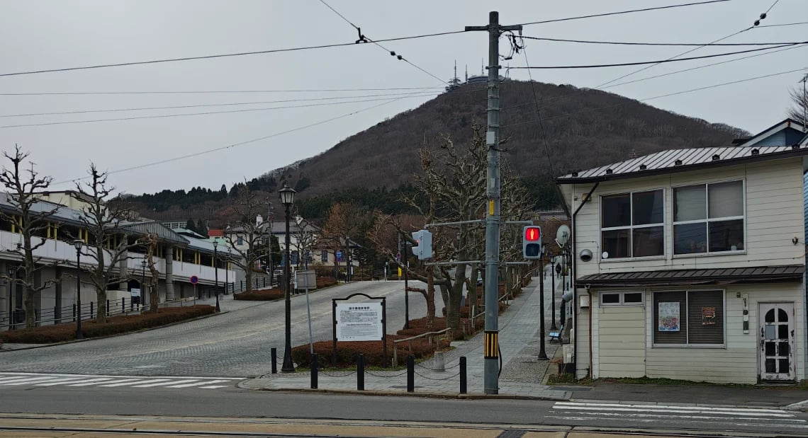A wide cobblestone street sloping upwards towards Mt. Hakodate, which has a tower on its peak. A red traffic light is visible on a utility pole, and there is a white wooden building on the right side of the street.