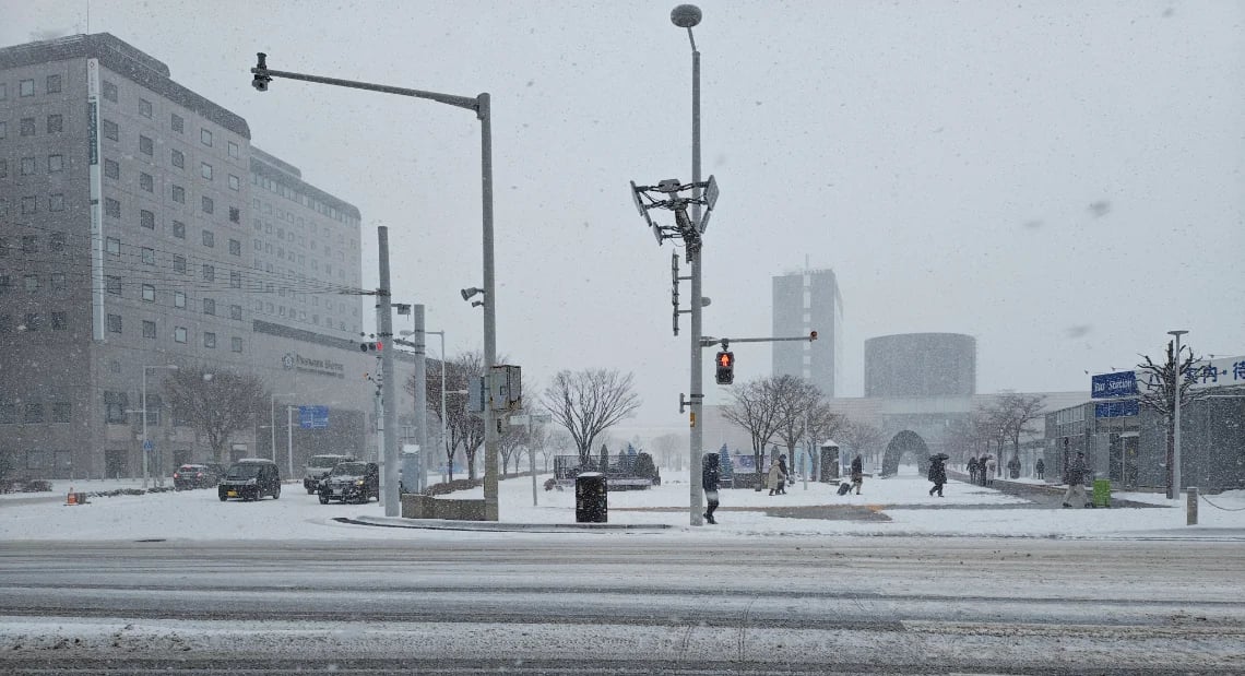 A snowy street scene at a large intersection in Hakodate during a heavy snowfall. The ground is covered in snow and slush, and pedestrians are walking on the sidewalks. A bus stop shelter is visible on the right, and a large hotel building is on the left.