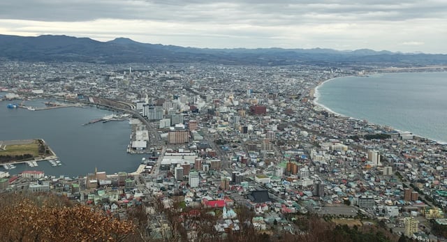 A panoramic view of the city of Hakodate from the summit of Mt. Hakodate on a cloudy day, showing the city's unique shape, the busy harbor on the left, and the open ocean on the right.