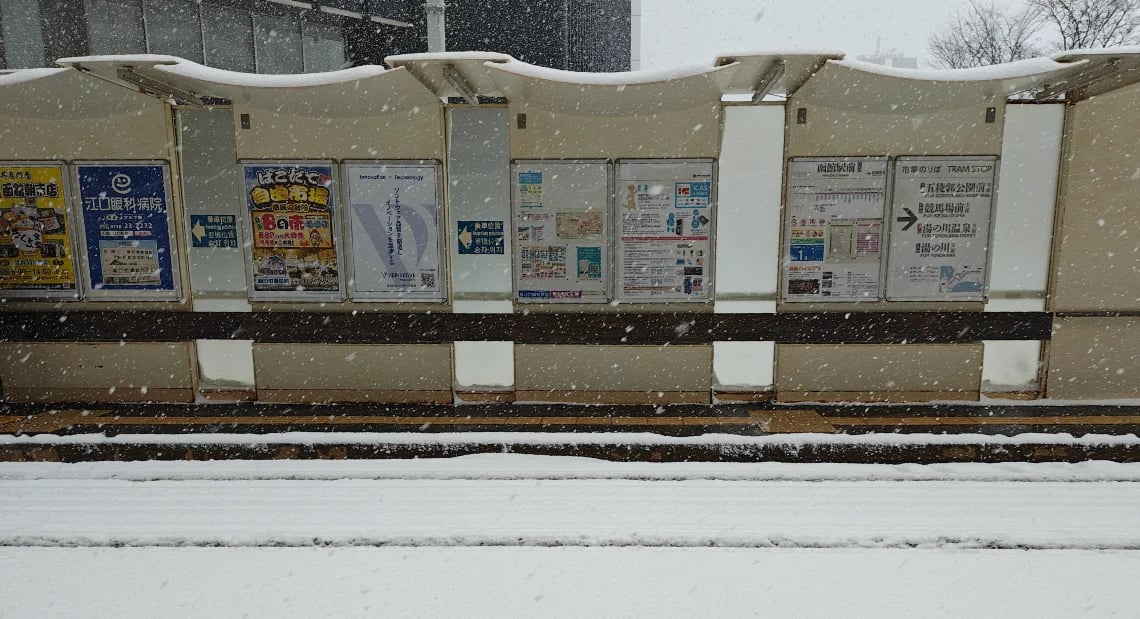A tram stop shelter in Hakodate covered in fresh snow while snow is falling. Timetables and advertisements are visible on the back panel of the shelter, and the tram tracks in the foreground are blanketed in white.