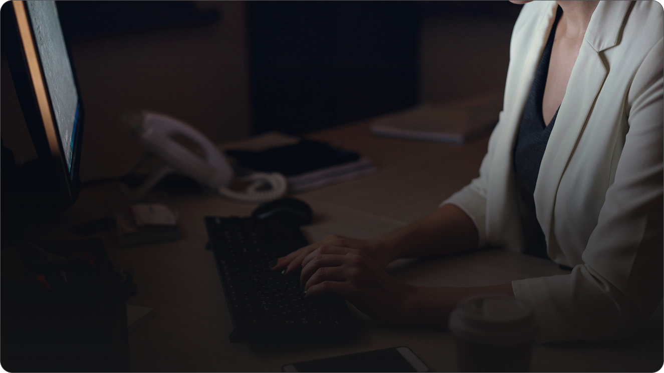 Close-up of a businesswoman in a white blazer typing on a keyboard at her desk in a dimly lit office, illuminated by the glow of her computer screen.