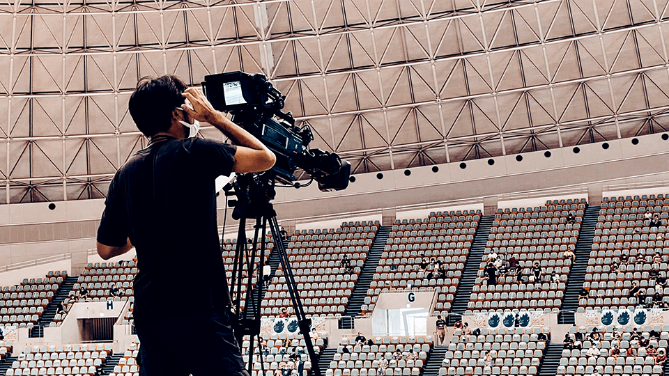 Broadcast camera operator filming from a tripod-mounted camera inside a stadium.