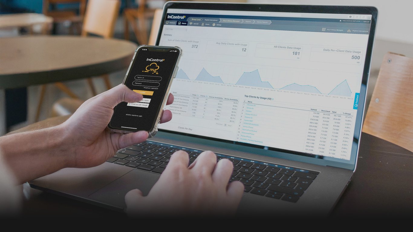Hand holding smartphone with Peplink InControl 2 app, laptop showing network dashboard in background.