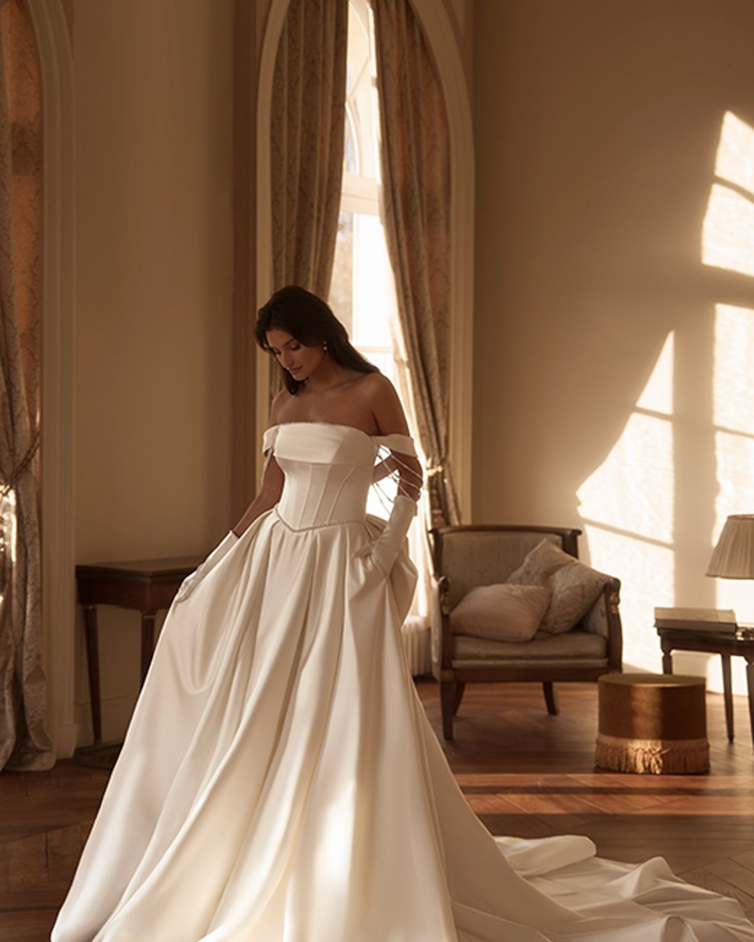 Model Hemina to klasyczna princesska w nowoczesnym wydaniu. Gładka satyna, prosty dekolt z opadającymi ramiączkami i zdobienia z pereł tworzą wyjątkowo elegancką kompozycję. Efektowny pas w literę V podkreśla talię, a szeroka spódnica z trenem i kokardą z tyłu dodaje bajkowego charakteru.