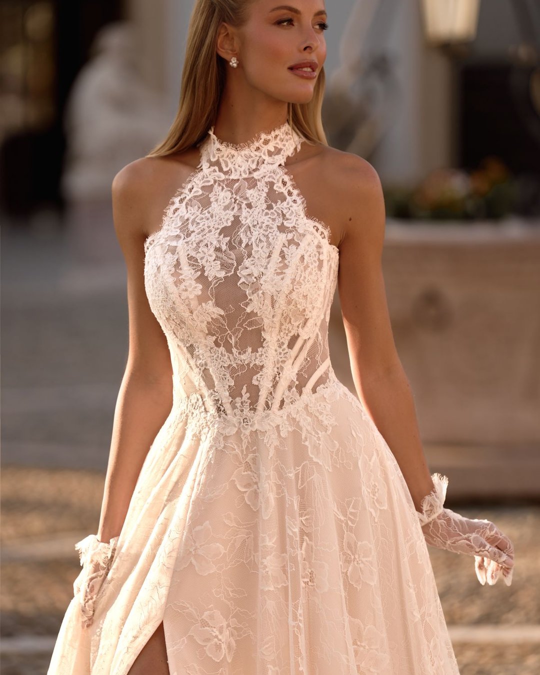 Suknia ślubna Ziggi od Anna Sposa to propozycja w stylu romantycznym, wykonana z delikatnej francuskiej koronki. Fason linii A, dekolt halter, wycięte plecy i rozporek nadają jej lekkości i nowoczesności. Subtelna przezroczystość, pasek w literę V oraz dopełniające stylizację koronkowe rękawiczki czynią tę kreację wyjątkowo zmysłową i elegancką.