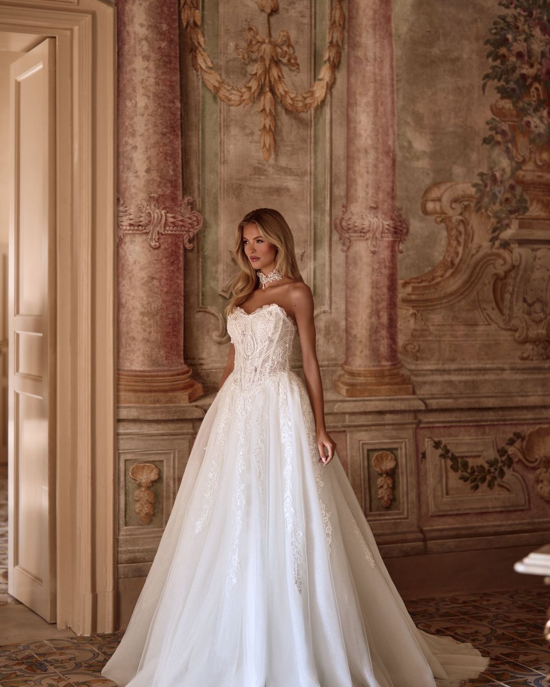 Karavella to zjawiskowa suknia ślubna w stylu princesski z efektowną szeroką spódnicą, wyrazistym gorsetem i delikatnym chokerem z perełkami. Zdobiona koronką i tiulem, zachwyca trenem oraz pięknym pasem w literę V, który podkreśla talię.