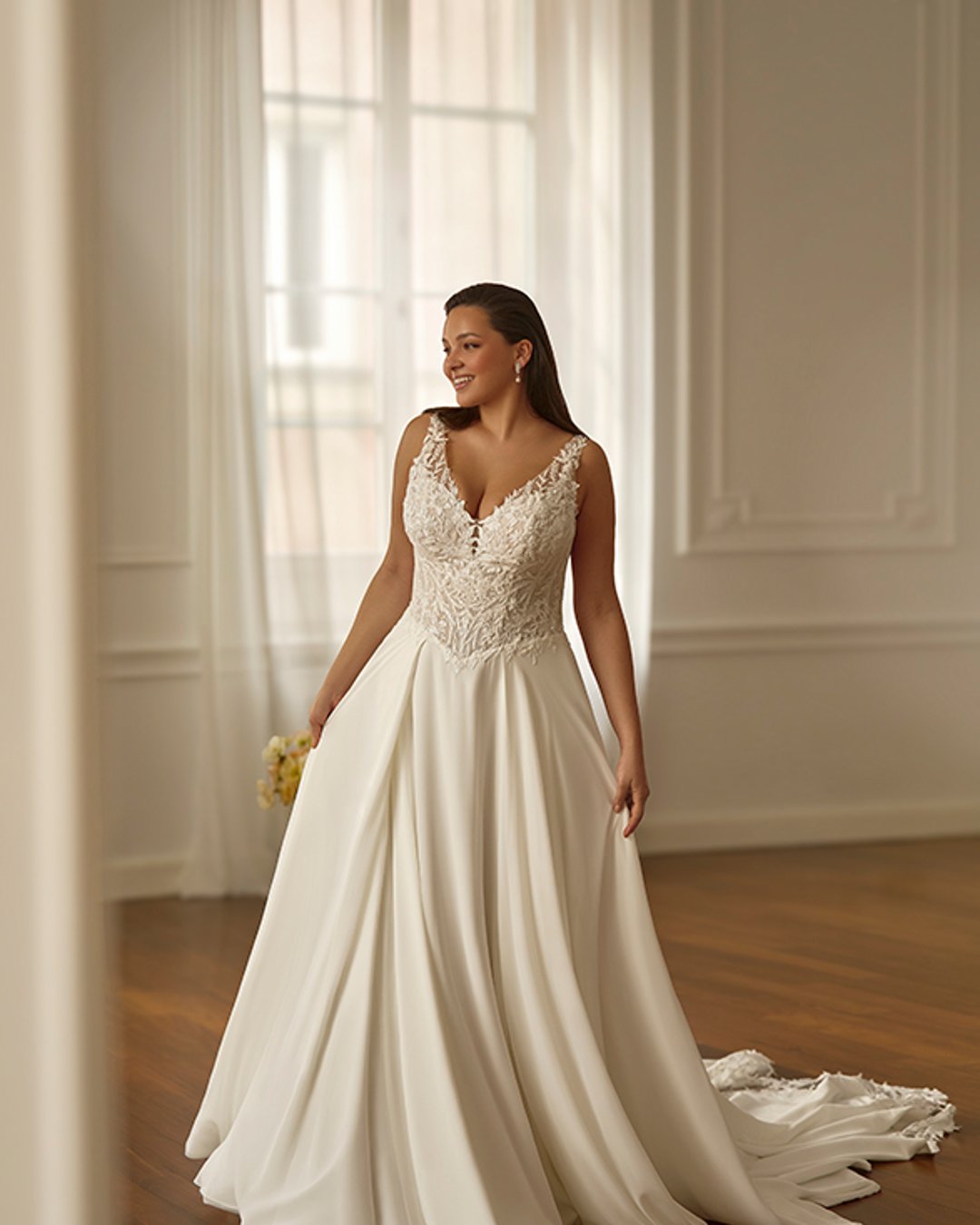 Model Shimella to klasyczna suknia ślubna o kroju linii A, z elegancką koronkową górą i zwiewną muślinową spódnicą. Przezroczysty gorset z szerokimi ramiączkami i pasem w literę V pięknie podkreśla talię i zapewnia komfort oraz subtelną elegancję.