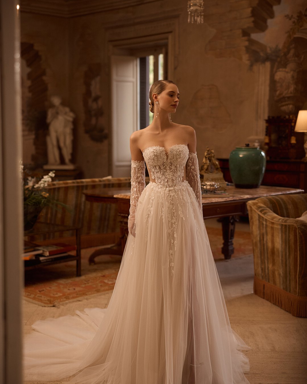 Suknia ślubna 5831 z kolekcji Elizabeth Passion łączy nowoczesny design z romantycznym wyrafinowaniem. Przezroczysta góra ozdobiona delikatną koronką i kwiatowymi aplikacjami, w połączeniu z szeroką, tiulową spódnicą, tworzy niepowtarzalny efekt, który zachwyca blaskiem. Nakładane rękawy dodają całości subtelności, sprawiając, że każda panna młoda poczuje się jak prawdziwa królewna w tym wyjątkowym dniu.