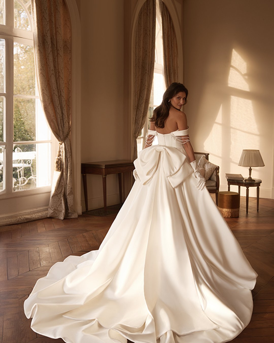 Model Hemina to klasyczna princesska w nowoczesnym wydaniu. Gładka satyna, prosty dekolt z opadającymi ramiączkami i zdobienia z pereł tworzą wyjątkowo elegancką kompozycję. Efektowny pas w literę V podkreśla talię, a szeroka spódnica z trenem i kokardą z tyłu dodaje bajkowego charakteru.