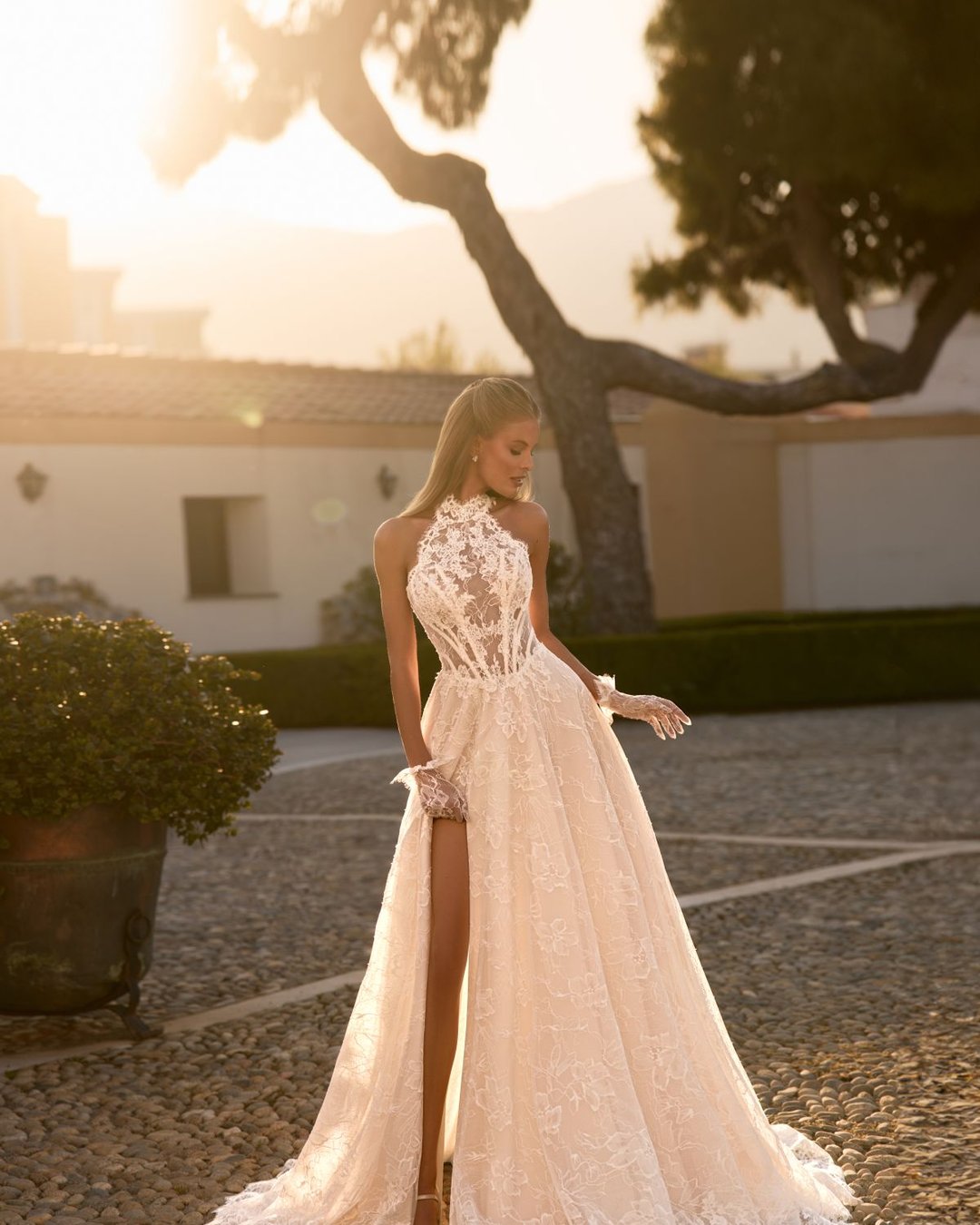 Suknia ślubna Ziggi od Anna Sposa to propozycja w stylu romantycznym, wykonana z delikatnej francuskiej koronki. Fason linii A, dekolt halter, wycięte plecy i rozporek nadają jej lekkości i nowoczesności. Subtelna przezroczystość, pasek w literę V oraz dopełniające stylizację koronkowe rękawiczki czynią tę kreację wyjątkowo zmysłową i elegancką.