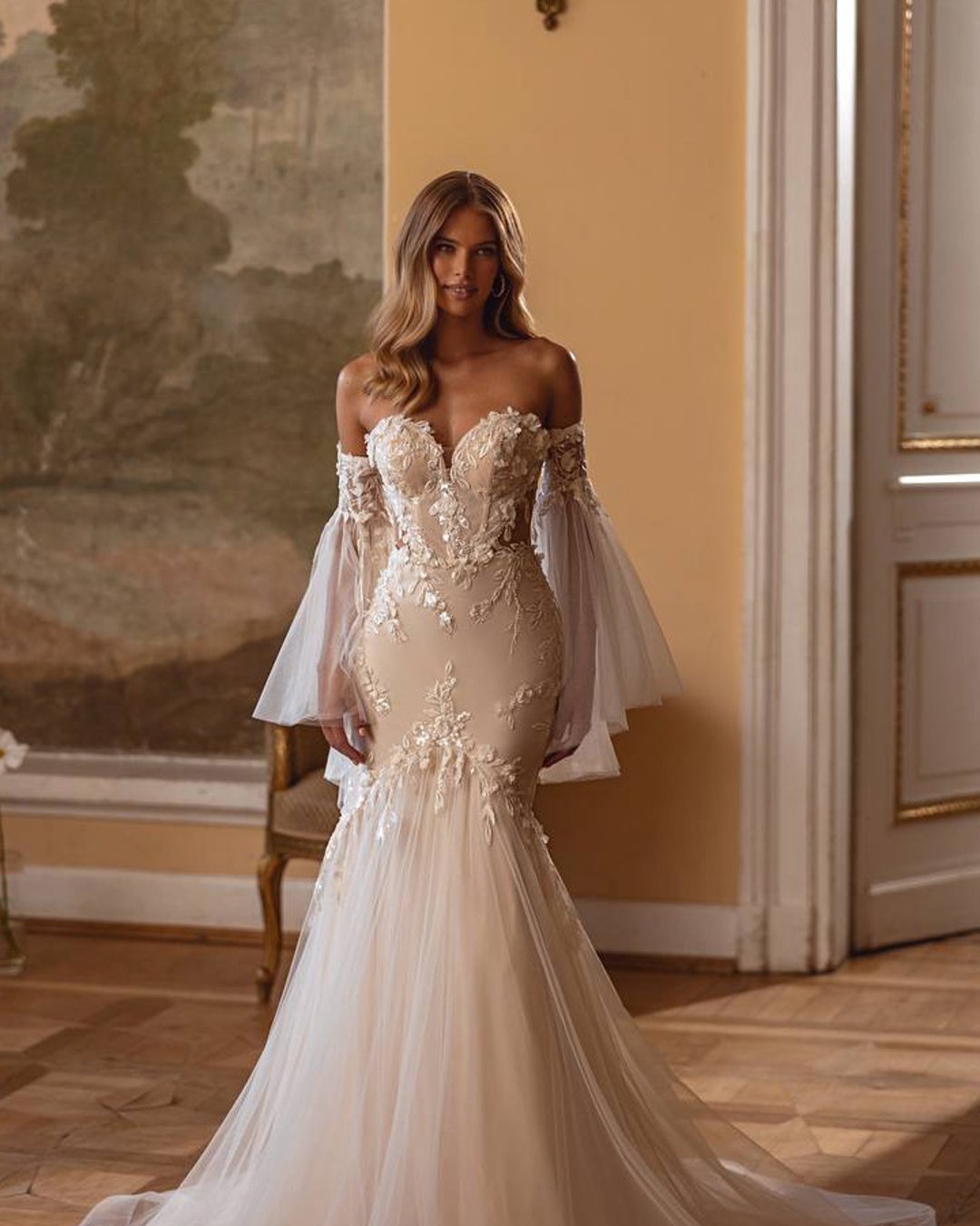 Suknia Bronte to połączenie klasycznej elegancji i nowoczesnej finezji. Wykonana z delikatnej koronki, idealnie dopasowuje się do sylwetki, a gorset ozdobiony misternymi aplikacjami kwiatowymi harmonizuje z opadającymi rękawami. Dolna część sukni rozszerza się, tworząc subtelny efekt ryby, a lekko opadający tiulowy tren nadaje całości majestatycznego wyglądu.