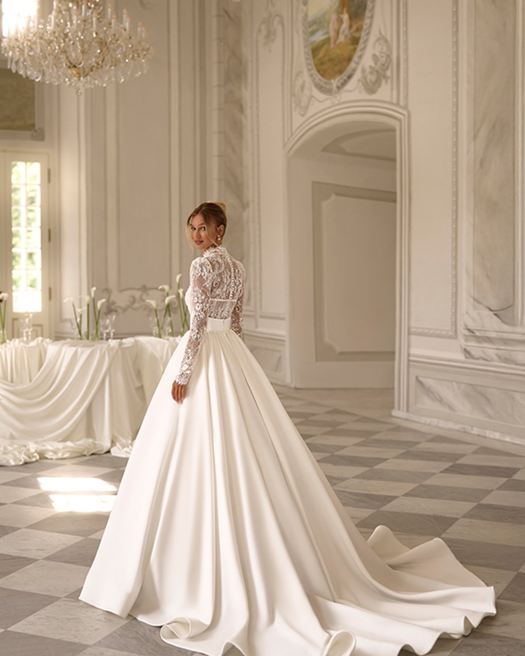 Calayra od Anna Sposa to elegancka suknia ślubna typu princessa, która zachwyca zmysłowym dekoltem w kształcie litery V, długimi koronkowymi rękawami oraz olśniewającą, rozkloszowaną satynową spódnicą z trenem. To połączenie klasyki i nowoczesnej delikatności.