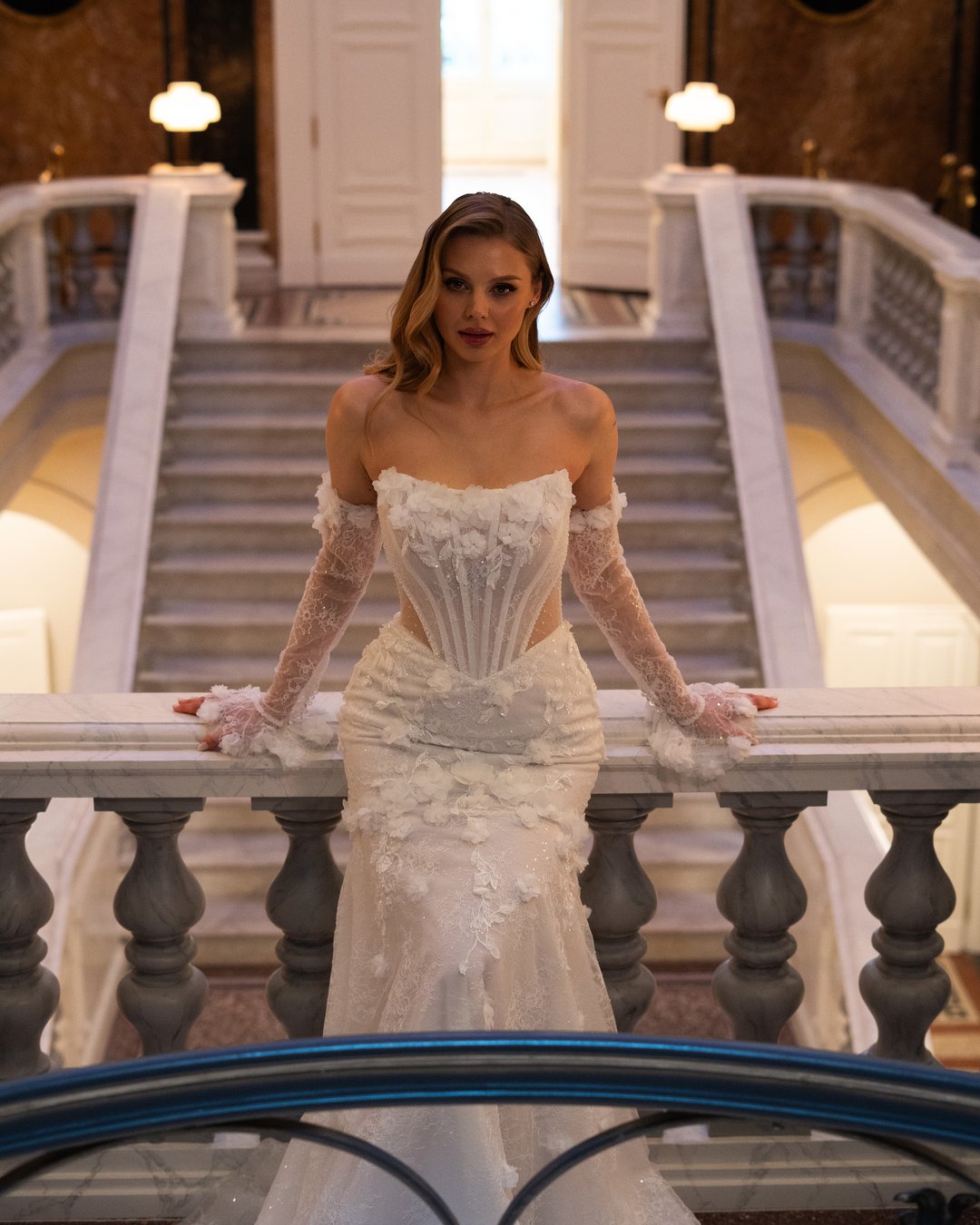 Suknia ślubna Charlotte z kolekcji LaLiven to prawdziwa ucieleśnienie elegancji i nowoczesnego romantyzmu. Jej koronkowy gorset z efektownym pasem w kształcie litery V oraz nakładane rękawy doskonale podkreślają sylwetkę, a błyszczące detale i kwiatowe motywy dodają subtelnej finezji. Model w stylu syrenki, idealnie dopasowany, sprawi, że każda panna młoda poczuje się wyjątkowo w swoim wielkim dniu.
