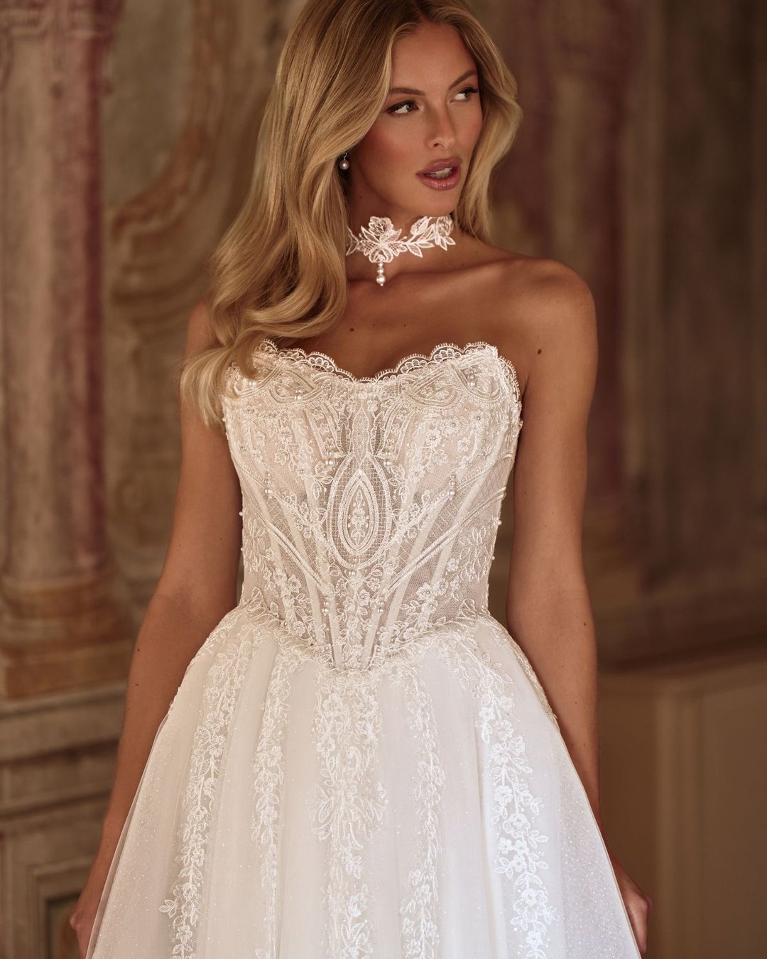 Karavella to zjawiskowa suknia ślubna w stylu princesski z efektowną szeroką spódnicą, wyrazistym gorsetem i delikatnym chokerem z perełkami. Zdobiona koronką i tiulem, zachwyca trenem oraz pięknym pasem w literę V, który podkreśla talię.