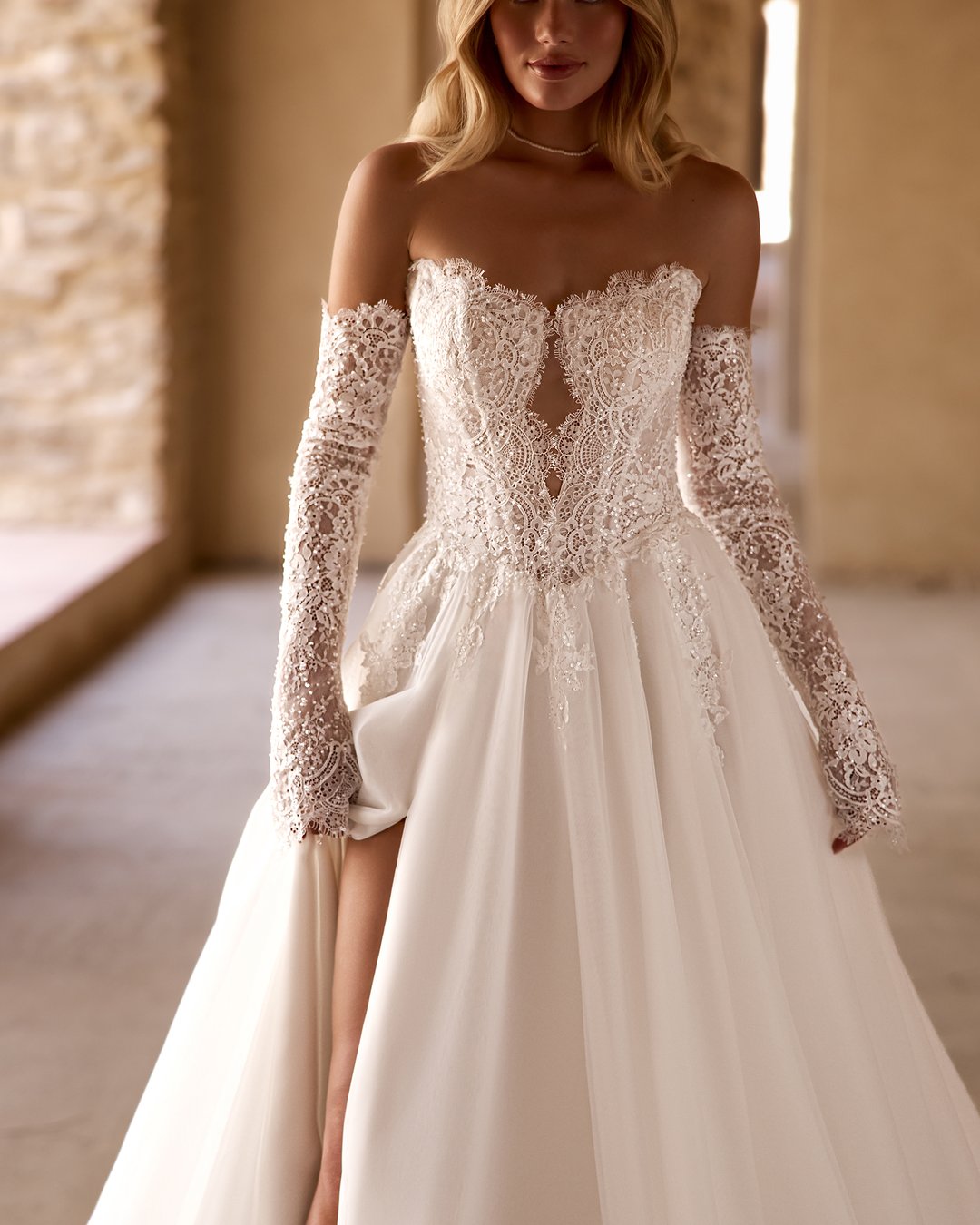 Suknia ślubna Forutina z kolekcji AnnaSposa to romantyczny wybór dla wyjątkowej panny młodej. Jej rozkloszowana linia A, zdobiona koronką francuską, zachwyca przezroczystymi detalami oraz efektownym trenem, natomiast gorsetowy krój z wyciętymi plecami podkreśla sylwetkę, a subtelny rozporek dodaje nuty zmysłowości. Dodatkowe, nakładane rękawy wprowadzają elegancję, sprawiając, że każda chwila w tej sukni będzie niezapomniana.