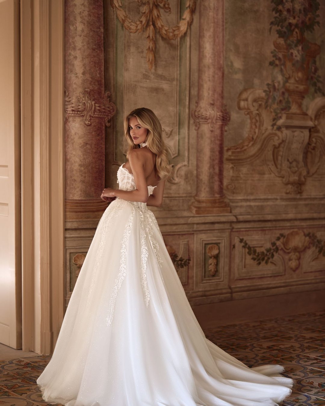 Karavella to zjawiskowa suknia ślubna w stylu princesski z efektowną szeroką spódnicą, wyrazistym gorsetem i delikatnym chokerem z perełkami. Zdobiona koronką i tiulem, zachwyca trenem oraz pięknym pasem w literę V, który podkreśla talię.