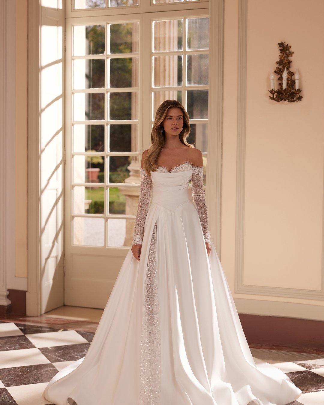 Suknia ślubna Vertalia z kolekcji AnnaSposa to kwintesencja klasycznej elegancji. Jej szeroka spódnica z rozcięciem i efektownym trenem w połączeniu z gorsetowym pasem w kształcie litery V oraz dekoltem serduszko podkreślają kobiecą sylwetkę, a delikatna francuska koronka dodaje całości subtelnego uroku. W tej sukni każda panna młoda poczuje się wyjątkowo w dniu swojego ślubu.