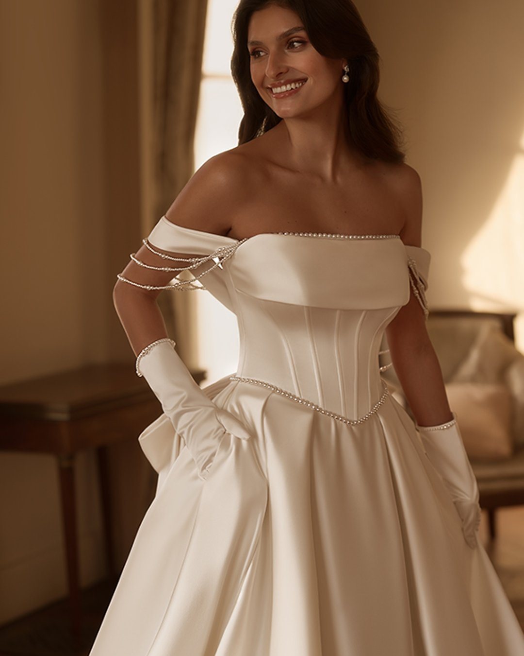 Model Hemina to klasyczna princesska w nowoczesnym wydaniu. Gładka satyna, prosty dekolt z opadającymi ramiączkami i zdobienia z pereł tworzą wyjątkowo elegancką kompozycję. Efektowny pas w literę V podkreśla talię, a szeroka spódnica z trenem i kokardą z tyłu dodaje bajkowego charakteru.