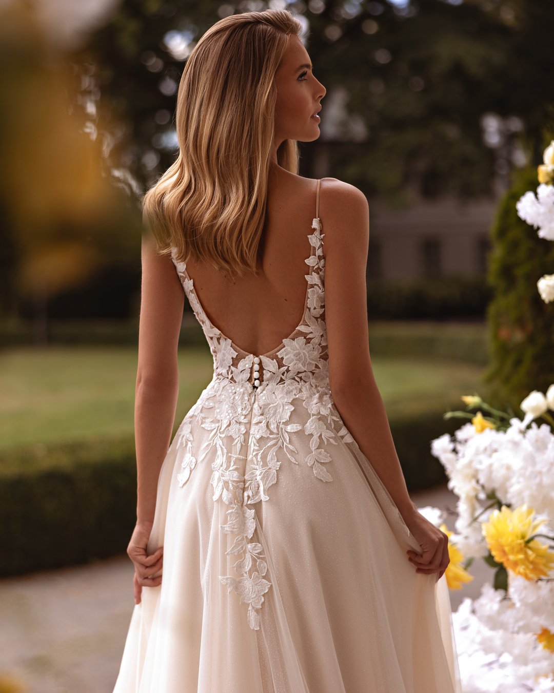 Suknia ślubna Kushni z kolekcji AnnaSposa to prawdziwa kwintesencja romantyzmu i elegancji. Odkryte plecy oraz szeroka spódnica w linii A, ozdobiona delikatną koronką i tiulem, sprawiają, że każda panna młoda poczuje się jak w bajce. Efektowny tren i subtelne ramiączka podkreślają wyjątkowość tej kreacji, a jej subtelny blask doda magii każdemu ślubnemu momentowi.